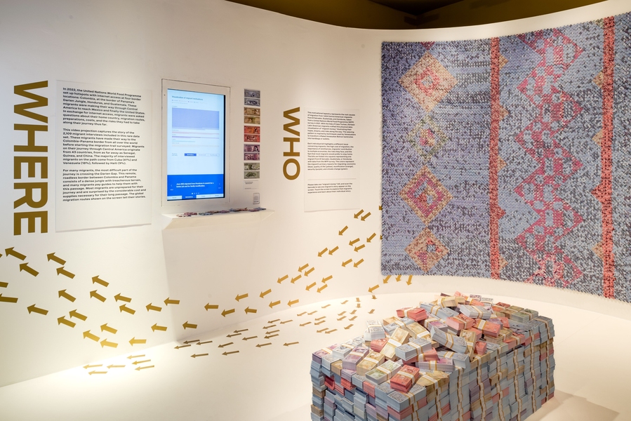 Art exhibit with wall and floor installations prominently features a large stack of bank notes