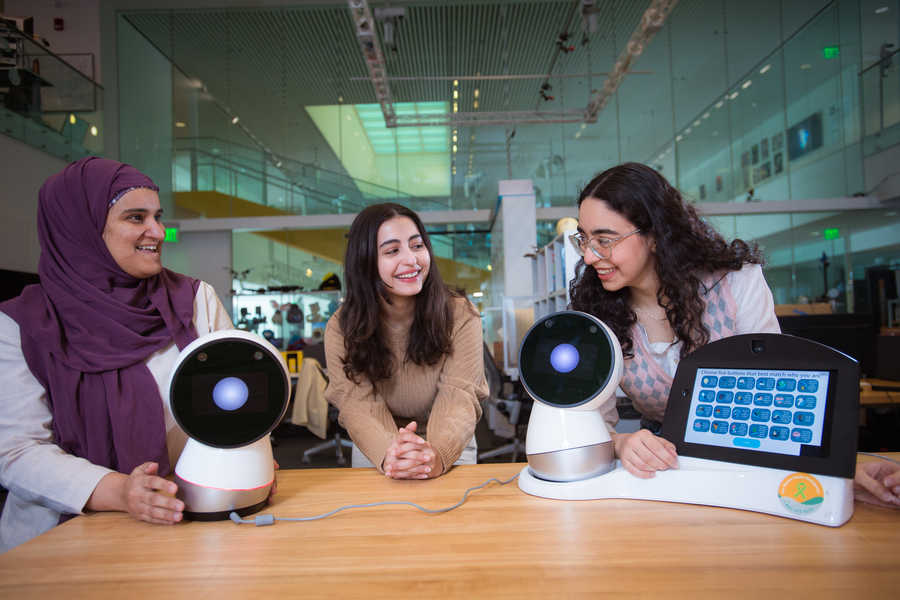 Sharifa Alghowinem with head scarf and Deim Alfozan and Tasneem Burghleh, both with long dark brown hair, lean on a table bearing two Jibo personal robots, which look like spheres with large, single eyes.