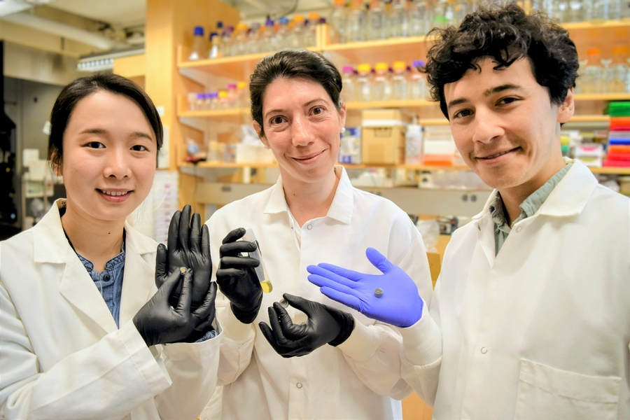 Qijun Liu, Maria Eugenia Inda, and Miguel Jimenez, all in lab coats and nitrile gloves, pose with prototypes of a pill-like capsule and a glass vial with yellow liquid