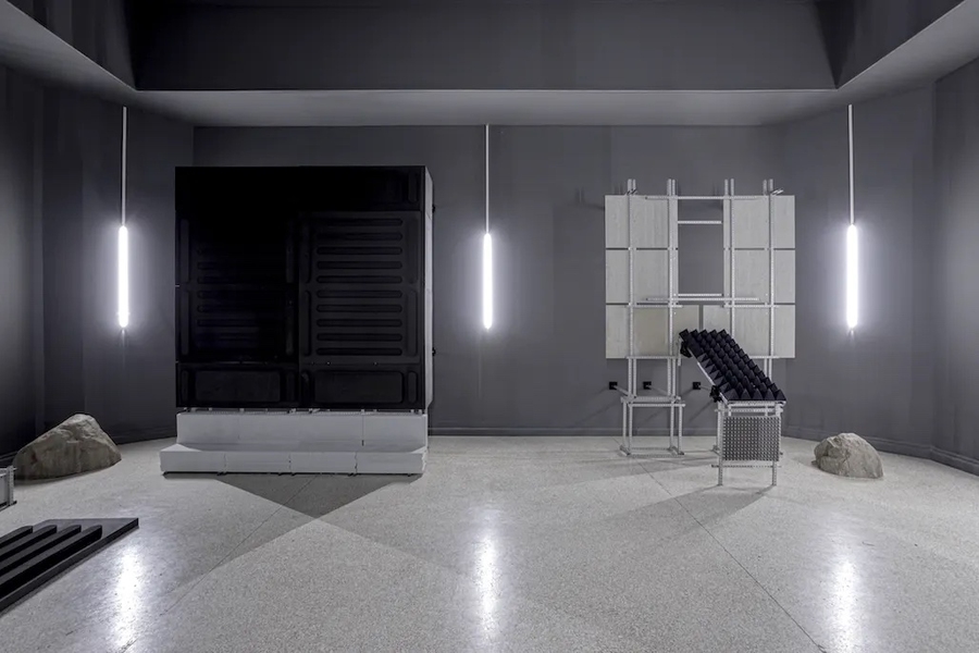 A series of furniture-like plastic objects in a monochrome gray room; two boulders are embedded in the floor.
