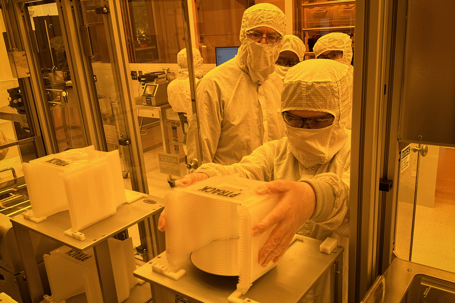 In a cleanroom suffused with amber-colored light, a person in a full bunny suit reaches into a large instrument holding a container of silicon wafers.