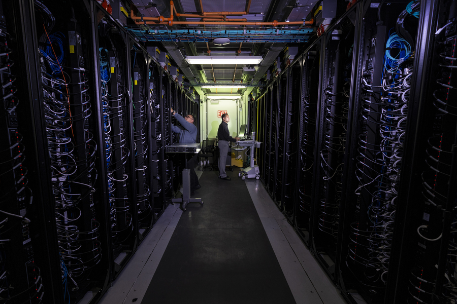 View of two researchers down a dark corridor in a data center, making adjustments to hardware on large racks lining both walls. 