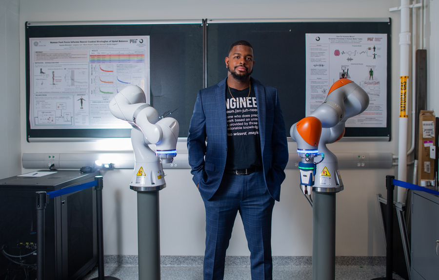 Michael West, wearing a suit with an "Engineer" T-shirt, stands in front of chalkboard, flanked by two robotic arms.