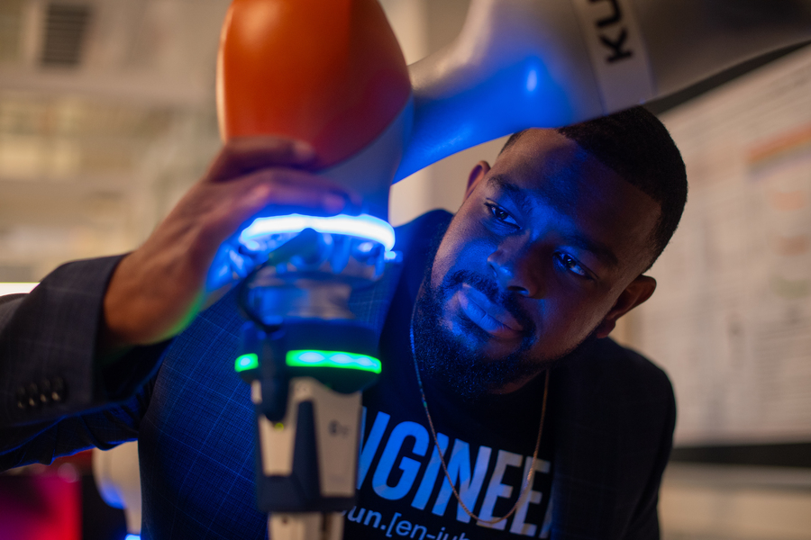 Close-up of a Michael West in an "engineer" T-shirt adjusting an orange robotic arm with colored lights casting a blue glow on his face