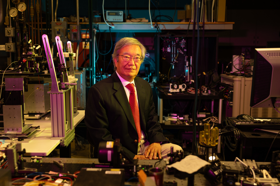 James Fujimoto sits in a crowded laboratory surrounded by imaging equipment. The lab is darkly lit with low fuschia, gold, and teal lights. 