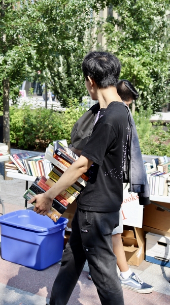 A man with a large stack of books in his hands walks by tables filled with books. 