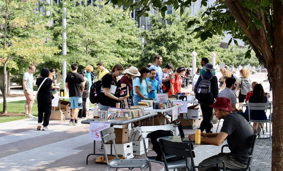 An outdoor area shaded by trees and two rows of tables filled with books is well-attended by shoppers.