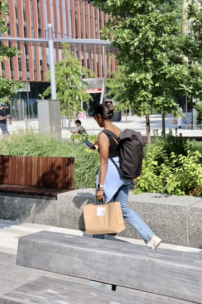 A woman wearing a backpack walks away from the camera, looking at her phone, with a brown paper bag in hand. 
