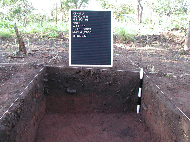 An archeological site is divided, by white rope, into a grid. One square has been dug out, and a sign says, “Xingu Nokugu… May 4, 2005. Midden”