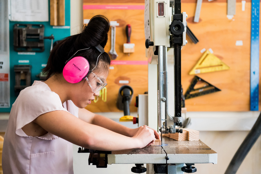 Photo of Trang L., who wears goggles and pink protective headphones, in profile using a bandsaw in a makerspace