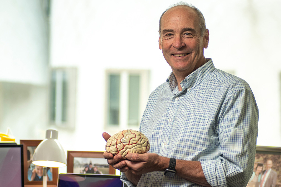 Mark Bear holds a model human brain. He poses in front of his desk, which feature family photos in frames, while the building next door is seen through glass windows behind him.