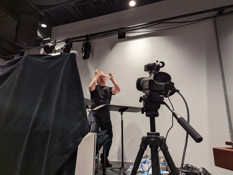 Tod Machover stands in front of a video camera on a rehearsal set, gesturing and speaking to someone off-camera