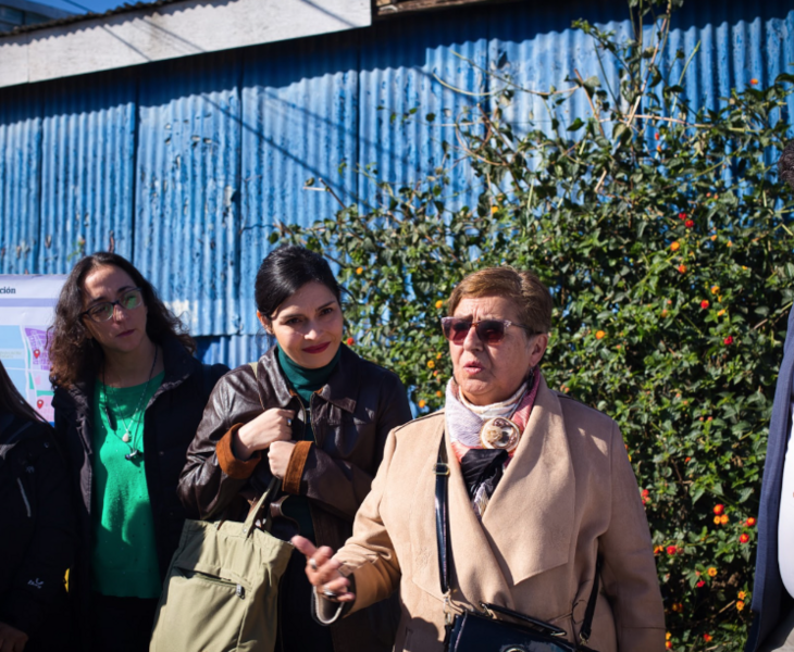 Katia Lobos, Marcela Martínez Bascuñán, and Rosa Terán stand outdoors in coats, in front of a flowering bush and a corrugated metal wall. 