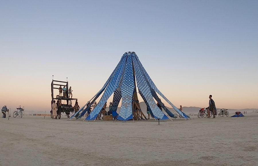 Sunset view of the Living Knitwork Pavilion, an open conical structure, in the desert. Sands stretch in front of the Pavilion, with mountains and sky in the distance.