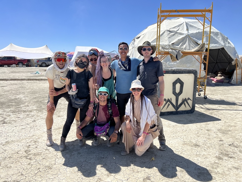 Eight people pose in the desert on a sunny day. A geometric dome tent appears behind them, with other tents visible even further in the background.