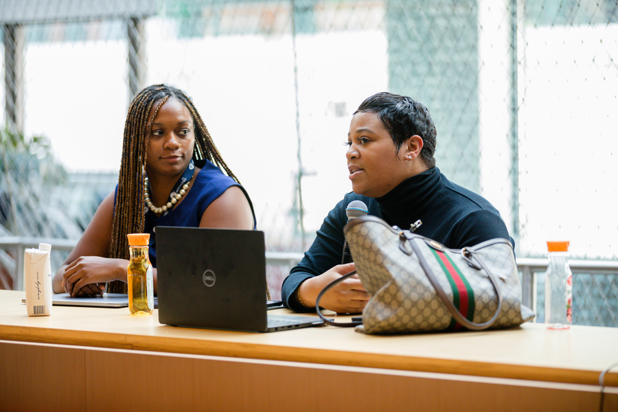 Two women sit at a table; one, with a laptop in front of her, is speaking into a microphone.