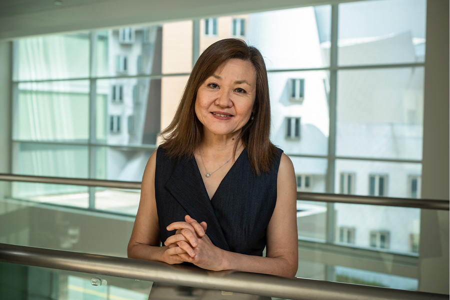  Li-Huei Tsai poses for a portrait, leaning against a metal railing. Brick and metal MIT buildings can be seen through a bank of windows behind her.