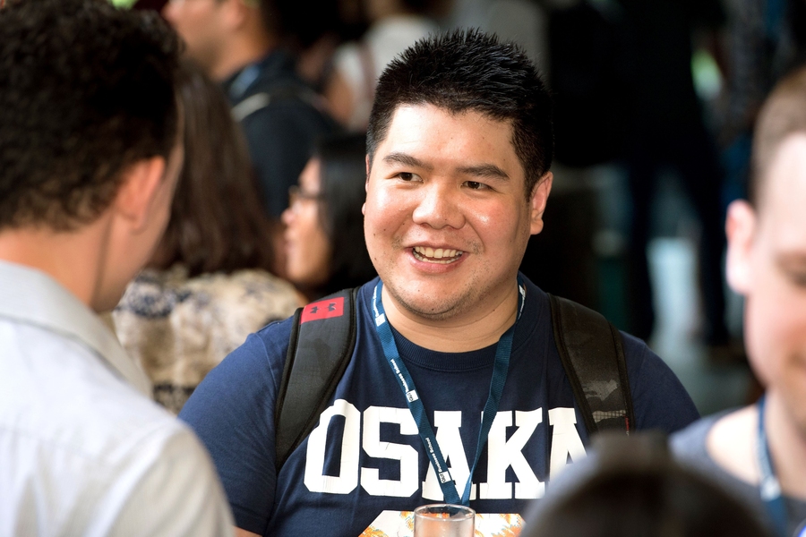 Nicky Agahari, wearing an Osaka T-shirt and a lanyard, speaking in a crowded room with a fellow bootcamper.