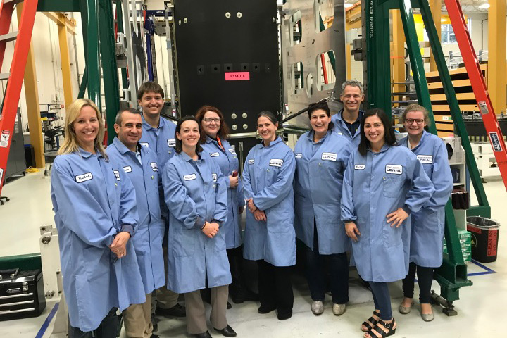 10 people in blue lab coats smile while inside a lab, with part of Psyche in background.