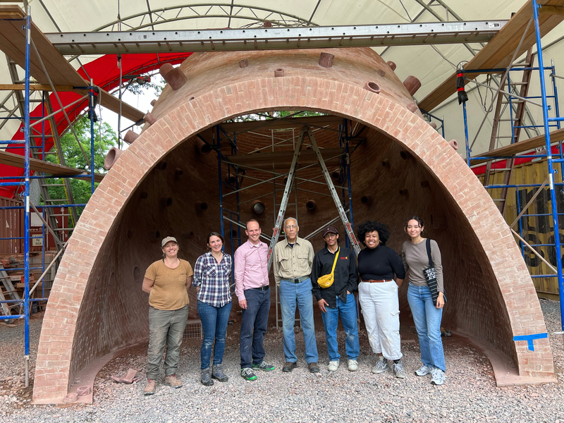 7 people stand inside the partially built structure, with ladders and equipment around the structure. From left to right: Lara Davis, Rebecca Buntrock, John Ochsendorf, Martin Puryear, Nebyu Haile, Nia Rich, and Domonique Valenzuela.