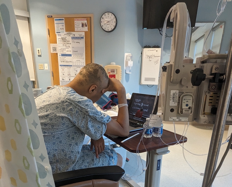 Dustin Liang seen in profile from slightly behind, sitting in front of his computer at a hospital. He is wearing a hospital gown and has lost most of his hair.