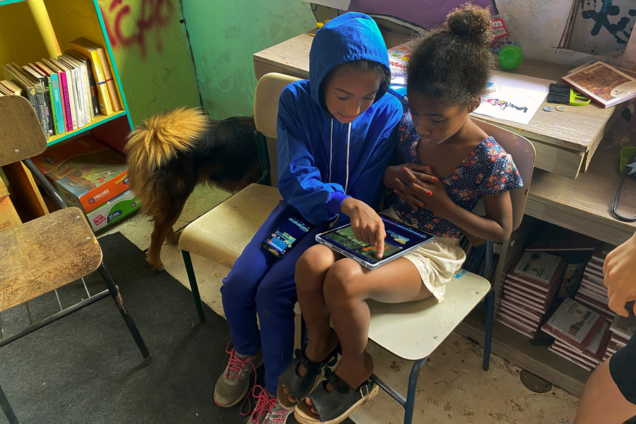 Two small children seated next to each other in a classroom share a tablet computer. There is a desk behind them and a bookshelf to the side.
