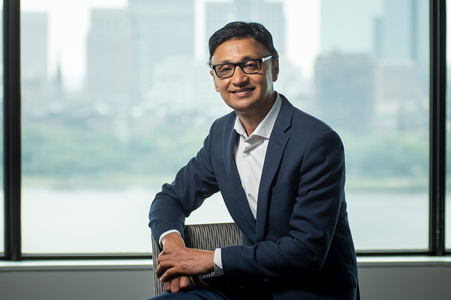 Rama Ramakrishnan posing sidesaddle in a chair with an out-of-focus skyline behind him