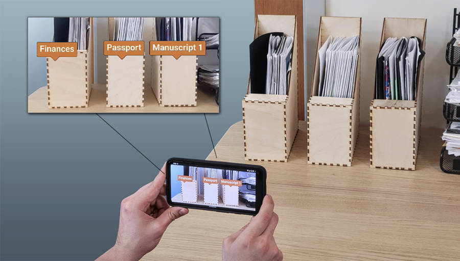 Scene of three small wooden file holders, while a hand holds a cellphone taking an image of the scene. On the phone's app, and in an enlarged inset, the file folders are labeled "Finances," "Passport," and "Manuscript 1"