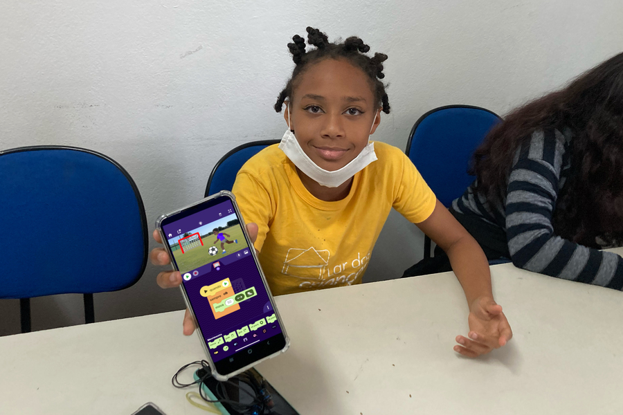 A child seated at a table in a white room holds a smartphone up to the camera to show off their soccer project.