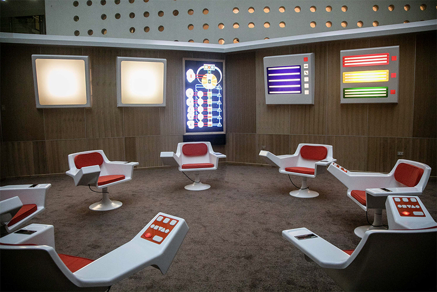 An empty, carpeted room with red-and-white chairs arranged in a circle and screens on the wall creating a futuristic appearance