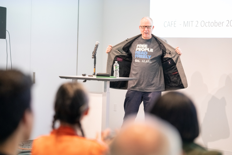 Richard Ovenden stands behind a lectern and opens his jacket to reveal a t-shirt that says "Free people read freely."