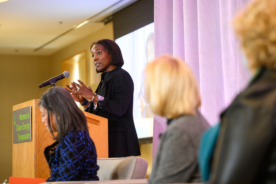 Paula Glover speaking and gesturing at a lectern with other panelists looking on in foreground.