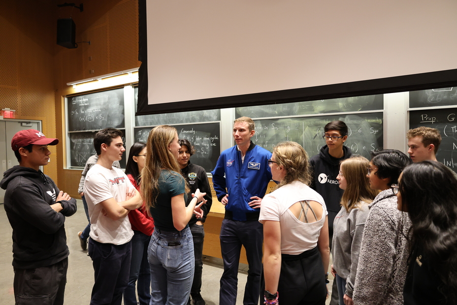 Woody Hoburg, wearing a blue NASA jacket, stands with hands on hips, talking to a dozen students in the front of a classroom
