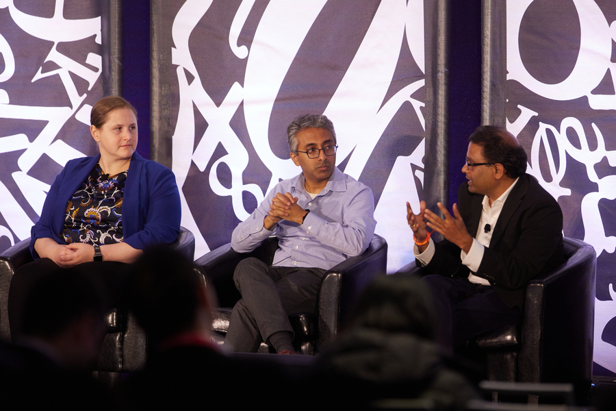 Three professors sit in chairs on a stage. Large white numbers and mathematical symbols appear behind them.