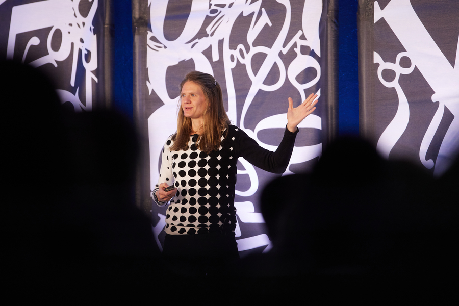 Seychelle Vos gestures with her left hand as she speaks from a stage. Large white numbers and mathematical symbols appear behind her.