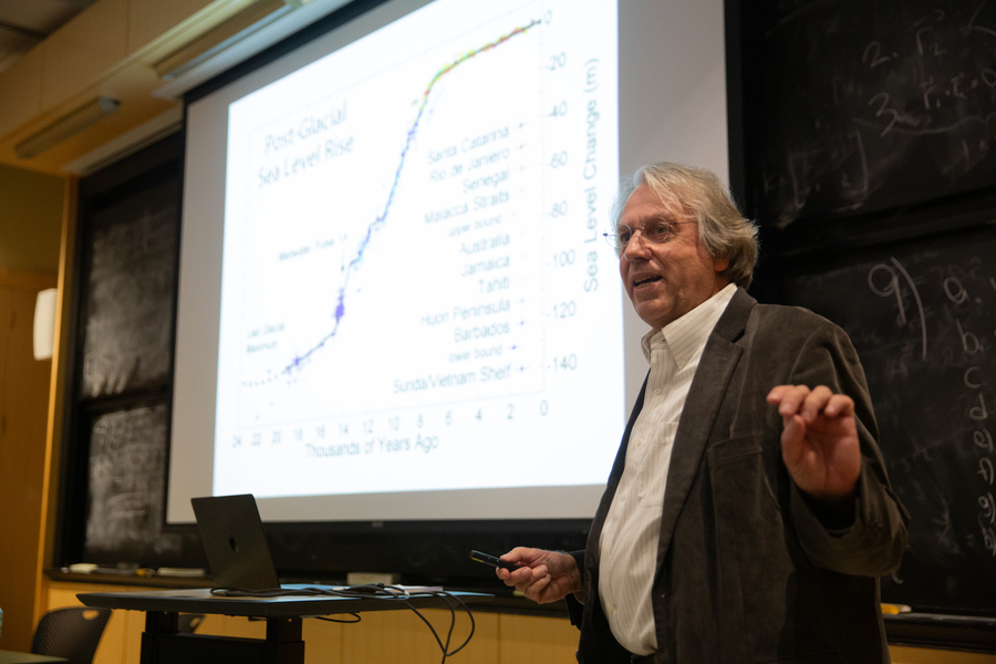 Kerry Emanuel stands in front of a classroom, addressing an audience which is out of frame. The screen behind him features an image plotting the course of sea level rise over millennia.