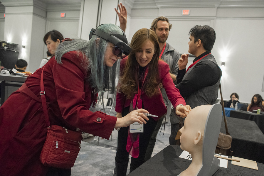 A woman points at a model of a human head while another woman with a VR headset reaches toward it with a small, pen-like device