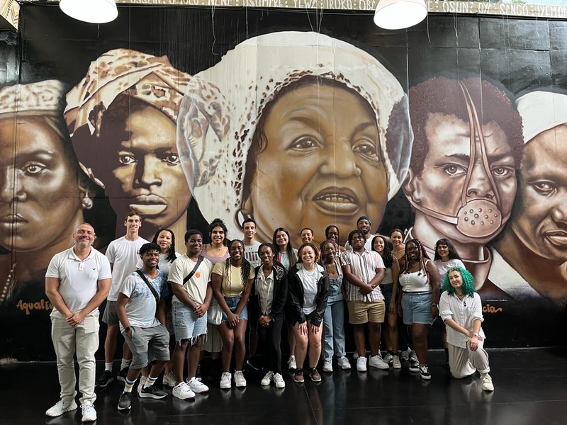 21 people pose in front of a mural depicting faces of five individuals of color