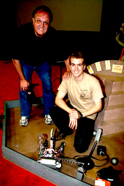 Randall Brings as a young boy kneels over a small robot while his father leans over him, hand on his shoulder.