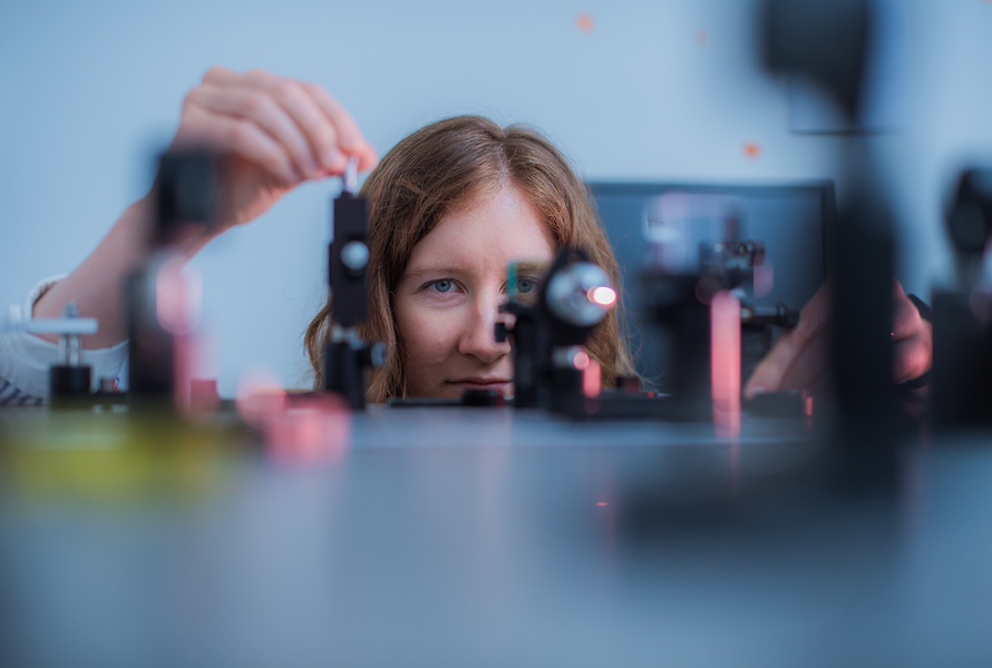 A crouching woman peers along the surface of a table of experiments with cameras and microscopes. Only her head from the mouth up is visible.