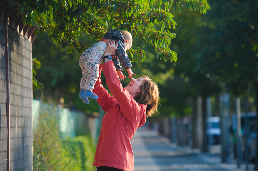 Woman holds baby above her head on a sidewalk with trees in background