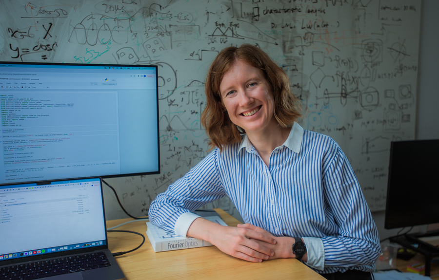Woman in blue striped shirt sits at desk near computers; white board with writing in background