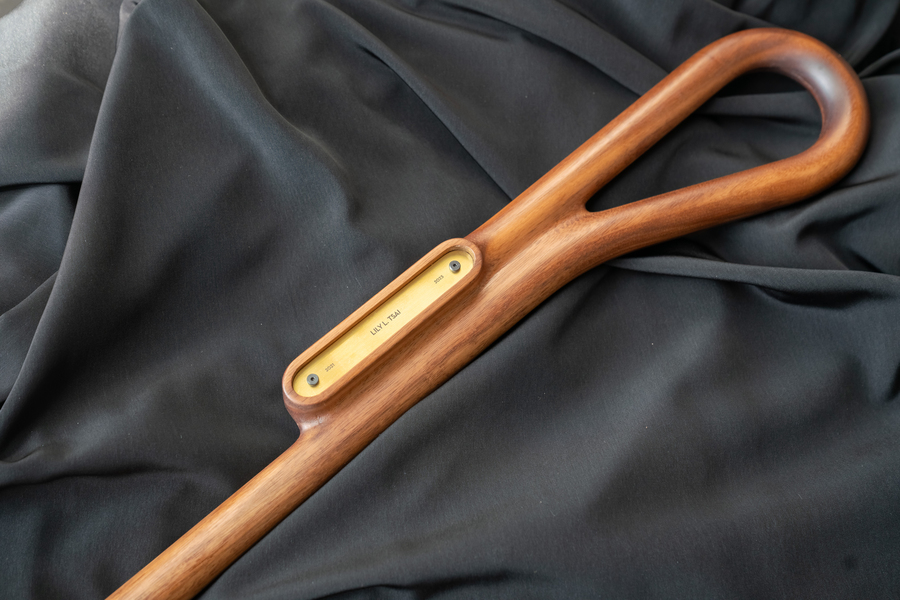 The curved handle of a wooden staff lying on a black cloth with an oblong open compartment containing a gold plate with a name and dates inscribed on it.