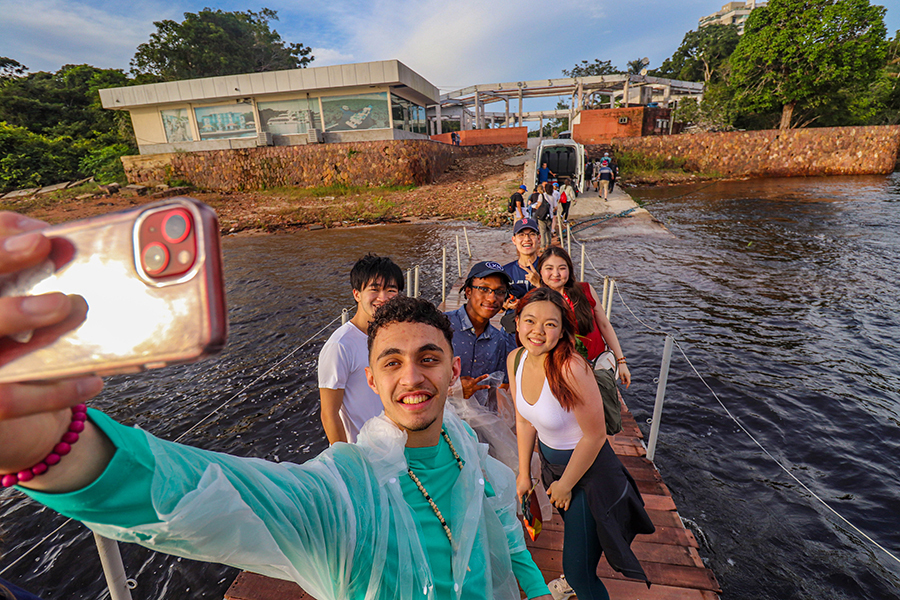 A student holds a camera, posing for a selfie with five others on a narrow boat on a river.
