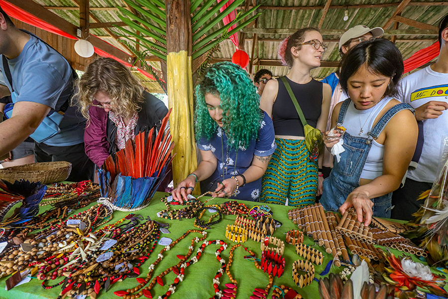 Three women bend over a table laden with native artwork and instruments in a bamboo hut in Brazil.