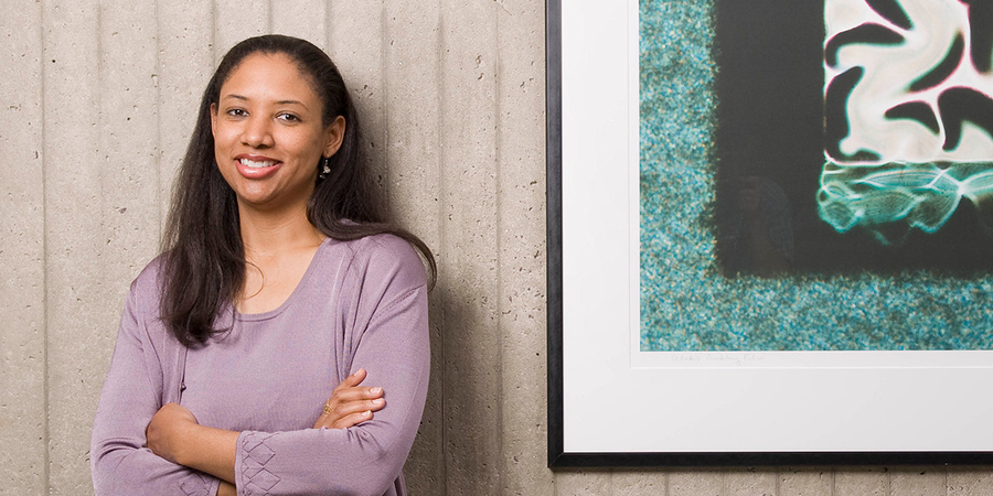 Kristala Prather poses for a photo in front of a cement wall, next to a framed green, black, and white illustration