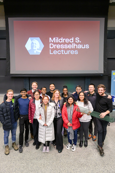 15 people stand in front of a screen that has "Mildred S. Dresslhaus Lecture" and a photo of Dresselhaus