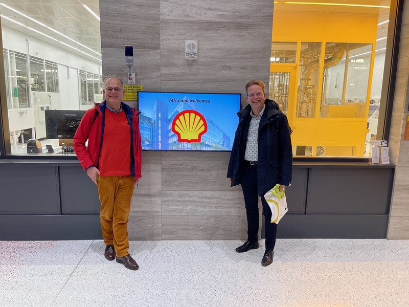 Alexander van der Made and Rolf van Benthem stand alongside a screen showing the logo for Shell in front of windows that look into the MIT.nano cleanroom laboratory.