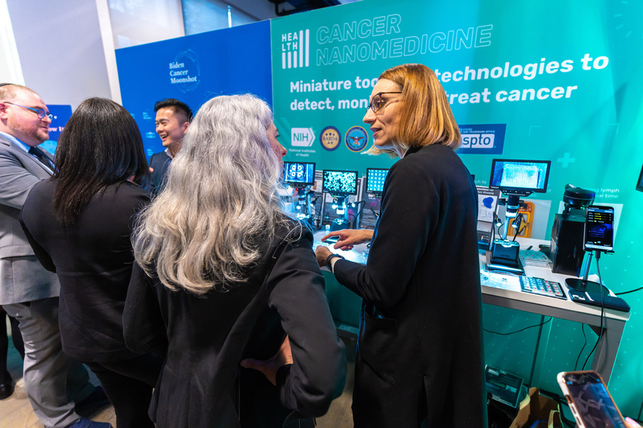 Ana Jaklenec speaks to a person with long silver hair with their back to the camera about the microscope screens on the table in front of them at a demonstration booth