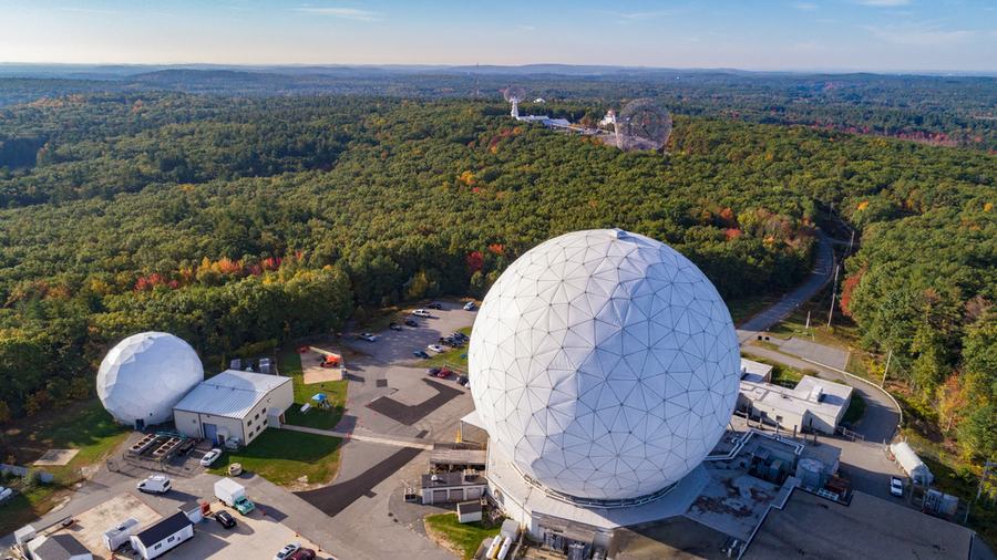 Aerial view of a large white geodesic dome, surrounded by small buildings and a large forest. Radio telescope dishes can be seen in the distance.